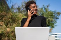 Mulher afro-americana em vestido preto elegante usando laptop e falando no telefone celular na rua — Fotografia de Stock