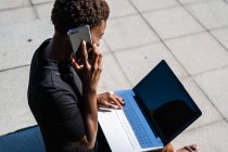 Афроамериканка в элегантном черном платье с ноутбуком и разговаривая по мобильному телефону на улице — стоковое фото