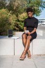 Концентрированная афроамериканка в элегантном черном платье с помощью мобильного телефона рядом с ноутбуком, расслабляясь на тротуаре — стоковое фото