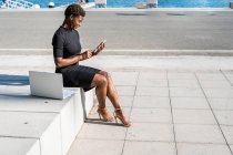 Вид сбоку сосредоточенной афроамериканки в элегантном черном платье, использующей мобильный телефон, сидя на тротуаре — стоковое фото
