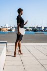 Mulher afro-americana em elegante vestido preto segurando laptop e usando telefone celular enquanto está em pé no pavimento — Fotografia de Stock