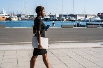 Femme afro-américaine en robe noire élégante tenant ordinateur portable et utilisant un téléphone portable tout en se tenant debout sur le trottoir — Photo de stock
