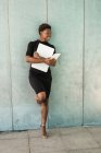 Coole afrikanisch-amerikanische intelligente Frau mit einem Handy, das einen Laptop hält, während sie an eine Betonwand gelehnt steht — Stockfoto