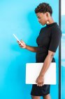 Mulher inteligente afro-americana legal usando telefone celular segurando laptop enquanto está em pé no fundo azul — Fotografia de Stock