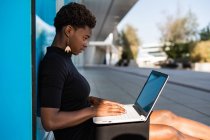 Side view of concentrated African American woman in elegant black dress using laptop while relaxing on pavement on street — Stock Photo