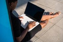 Concentrated African American woman in elegant black dress using laptop while relaxing on pavement on street — Stock Photo