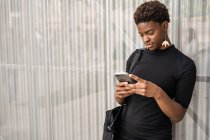 Mujer afroamericana con estilo enfocado en un teléfono inteligente de mensajería de vestido negro mientras está de pie sobre fondo metálico - foto de stock