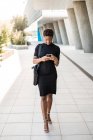 Обнаженная стильная афроамериканка в черном платье со смартфоном во время прогулки по улице — стоковое фото