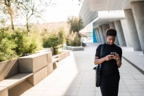 Enfocado mujer afroamericana con estilo en el teléfono inteligente de mensajería vestido negro mientras camina por la calle - foto de stock