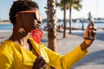 Модная афроамериканка в жёлтой куртке с леденцом у деревянного забора делает селфи с мобильным телефоном — стоковое фото