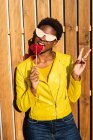 Mulher afro-americana na moda desfrutando de pirulito em forma de coração por cerca de madeira e sinal de paz gestual — Fotografia de Stock