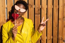 Trendy African American woman enjoying heart shaped lollipop by wooden fence and gesturing peace sign — Stock Photo