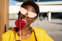 Trendy African American woman in sunglasses in yellow jacket enjoying heart shaped lollipop — Stock Photo