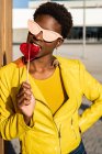 Mulher afro-americana na moda em óculos de sol em casaco amarelo desfrutando de pirulito em forma de coração por cerca de madeira — Fotografia de Stock