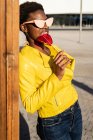 Mulher afro-americana na moda em óculos de sol em casaco amarelo desfrutando de pirulito em forma de coração inclinado por cerca de madeira — Fotografia de Stock