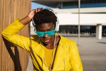 African American woman in stylish bright jacket and bright blue sunglasses using headphones standing near a modern building — Stock Photo