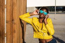 Mulher afro-americana em elegante casaco brilhante e óculos de sol azul brilhante usando fones de ouvido em pé perto de um edifício moderno — Fotografia de Stock