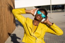 Donna afroamericana in elegante giacca luminosa e occhiali da sole blu brillante utilizzando cuffie in piedi vicino a un edificio moderno — Foto stock