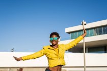 Baixo ângulo de mulher feliz afro-americana na elegante jaqueta brilhante e óculos de sol pulando com as mãos para cima — Fotografia de Stock