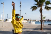 Bajo ángulo de la mujer afroamericana feliz en chaqueta brillante con estilo y gafas de sol saltando con las manos en la calle - foto de stock