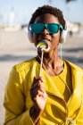 Mulher afro-americana em óculos de sol em casaco amarelo desfrutando de um pirulito e ouvindo música em fones de ouvido — Fotografia de Stock