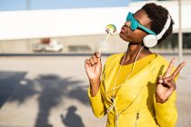 African American woman in sunglasses in yellow jacket enjoying a lollipop and listening to music on headphones — Stock Photo
