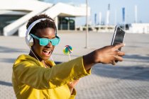 Donna afroamericana godendo lecca-lecca e ascoltando musica sulle cuffie mentre prende un selfie su un telefono cellulare — Foto stock