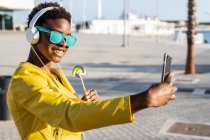 Femme afro-américaine appréciant la sucette et écoutant de la musique sur un casque tout en prenant un selfie sur un téléphone portable — Photo de stock