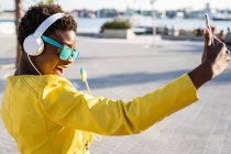 Афроамериканка наслаждается леденцом и слушает музыку в наушниках, делая селфи на мобильном телефоне — стоковое фото