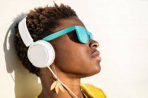 Afrikanisch-amerikanische Frau in stylischer heller Jacke und hellblauer Sonnenbrille mit Kopfhörern, die sich an eine weiße Wand lehnen — Stockfoto