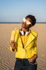 Trendy African American woman in headphones and sunglasses in bright yellow jacket enjoying ice cream in desert — Stock Photo
