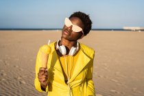 Mujer afroamericana de moda en gafas de sol con chaqueta de color amarillo brillante disfrutando de helado de pie en la playa de arena - foto de stock