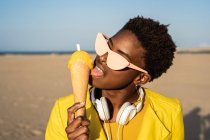 Trendige Afroamerikanerin mit Sonnenbrille in leuchtend gelber Jacke genießt Eis am Sandstrand — Stockfoto