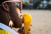 Вид сбоку на трепещущую афроамериканку в ярко-желтой куртке, наслаждающуюся мороженым, стоя на песчаном пляже — стоковое фото