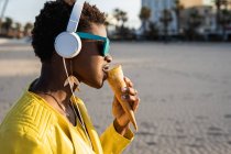 Side view of trendy African American woman in bright yellow jacket enjoying ice cream standing in sandy beach — Stock Photo