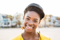 Portrait of happy African American woman in stylish bright jacket looking in camera on sandy beach blurred background — Stock Photo