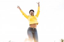 Low angle of happy African American woman in stylish bright jacket jumping with hands up on white background — Stock Photo
