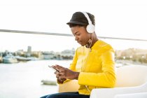 Seitenansicht einer Afroamerikanerin in stylischer heller Jacke, die ihr Handy benutzt und über Kopfhörer Musik hört — Stockfoto