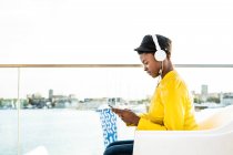 Vue latérale de la femme afro-américaine dans une veste lumineuse élégante utilisant un téléphone portable et écoutant de la musique sur écouteurs — Photo de stock
