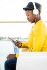 Vista lateral da mulher afro-americana em elegante casaco brilhante usando telefone celular e ouvir música em fones de ouvido — Fotografia de Stock