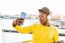 Mujer afroamericana con elegante chaqueta brillante tomando selfie y escuchando música en los auriculares - foto de stock