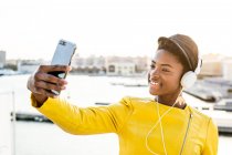 Афроамериканка в стильной яркой куртке делает селфи и слушает музыку в наушниках — стоковое фото