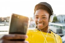Femme afro-américaine dans une veste lumineuse élégante prenant selfie et écouter de la musique sur écouteurs — Photo de stock