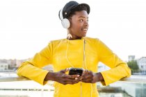 Donna afroamericana in elegante giacca luminosa utilizzando il telefono cellulare e ascoltando musica sulle cuffie — Foto stock