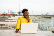 Tired African American woman in yellow jacket yawning while using laptop at wooden desk in city on blurred background — Stock Photo