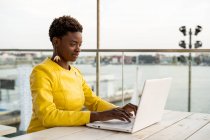 Mulher afro-americana em casaco amarelo usando laptop em mesa de madeira na cidade em fundo embaçado — Fotografia de Stock