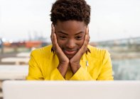 Surprised African American woman in yellow jacket using laptop at wooden desk in city on blurred background — Stock Photo