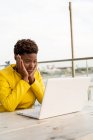 Rosto surpreso de mulher negra afro-americana em casaco amarelo usando laptop em mesa de madeira na cidade em fundo borrado — Fotografia de Stock