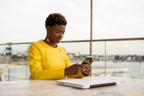 Smartphone de messagerie féminine afro-américaine concentré tout en se relaxant à la table en bois — Photo de stock