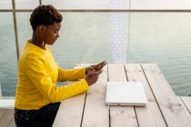 Mensagens femininas afro-americanas concentradas com smartphone enquanto relaxa na mesa de madeira — Fotografia de Stock
